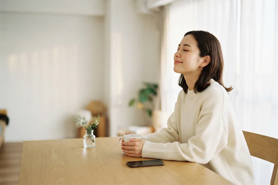 片付いたダイニングテーブルで、スマートフォンを伏せてマグを両手に持ち一息つく人